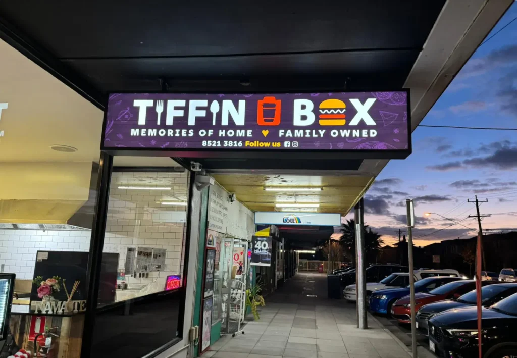Tiffin Box: Your Favourite Casual Burger Restaurant in Clayton 2 A storefront at dusk showcases a sign reading Tiffin Box adorned with icons of a tiffin and a burger. The sign whispers Memories of Home and Family Owned, just like the comforting welcome youd find on a homepage. Several parked cars rest beneath the partly cloudy sky, adding to the charm.