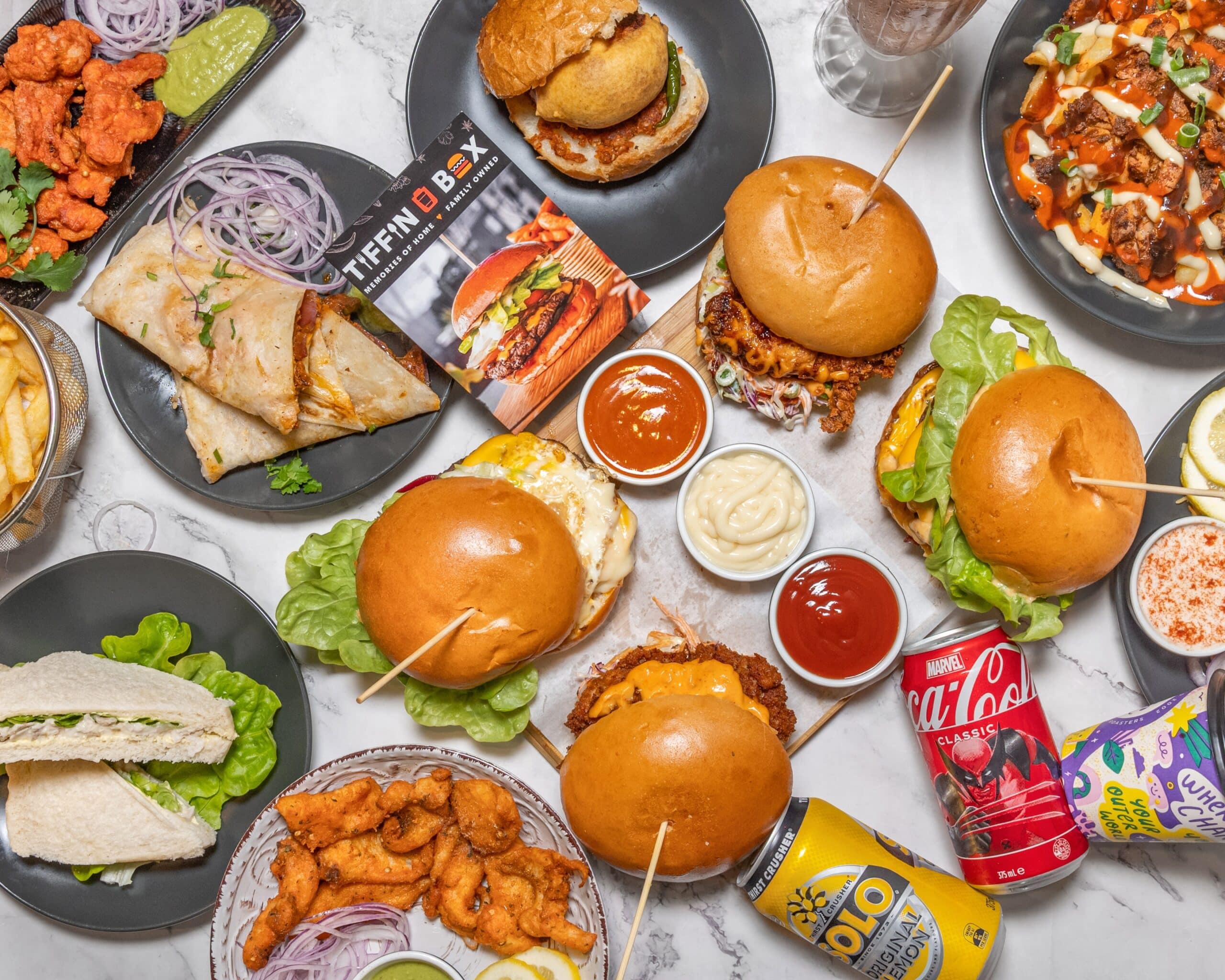 Find the Best Indian Burger – Clayton 3 An overhead view of a table brimming with delicious foods, including burgers, wraps, fries, and chicken wings, accompanied by cans of soda and a milkshake. Plates encircle a book titled THEMBOX. A chic marble table enhances the scenes elegance—a perfect spot to enjoy an Indian burger.