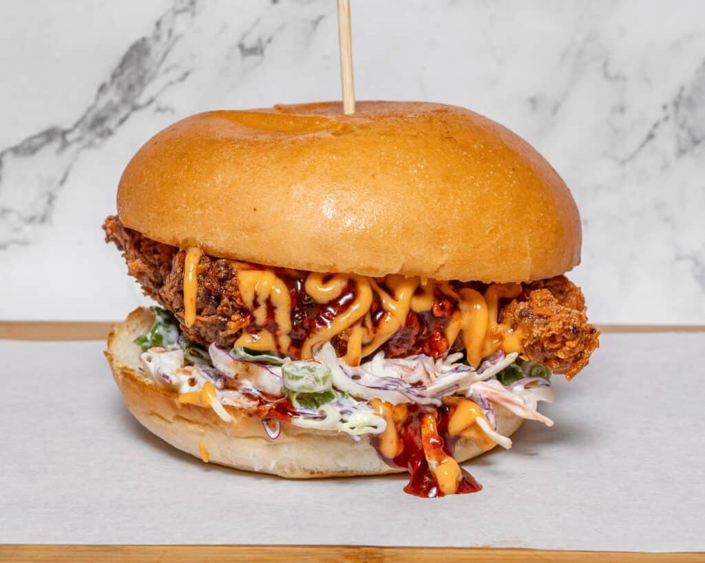 Top Rated Burger – Discover Melbourne’s Best Indian-Style Burgers at Tiffin Box 2 On a marble background, this top-rated burger near me features a crispy chicken fillet, creamy coleslaw, and spicy sauce, all nestled within a glossy, lightly toasted bun. A toothpick artfully secures the delicious creation on its wooden board.