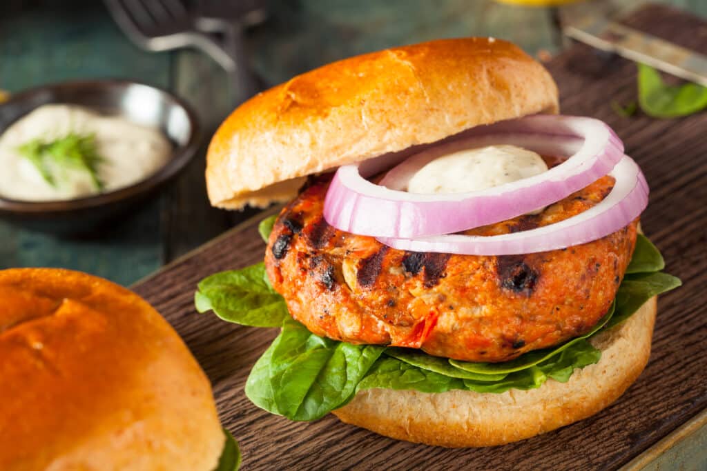 The Best Asian Burgers in Clayton You Need to Try Right Now 2 A grilled veggie burger on a bun with fresh spinach, topped with red onion slices and a dollop of sauce, served on a wooden board. Discover the flavors of Asian burgers in Clayton as a partial view of another bun and a small dish of sauce enhances the background.